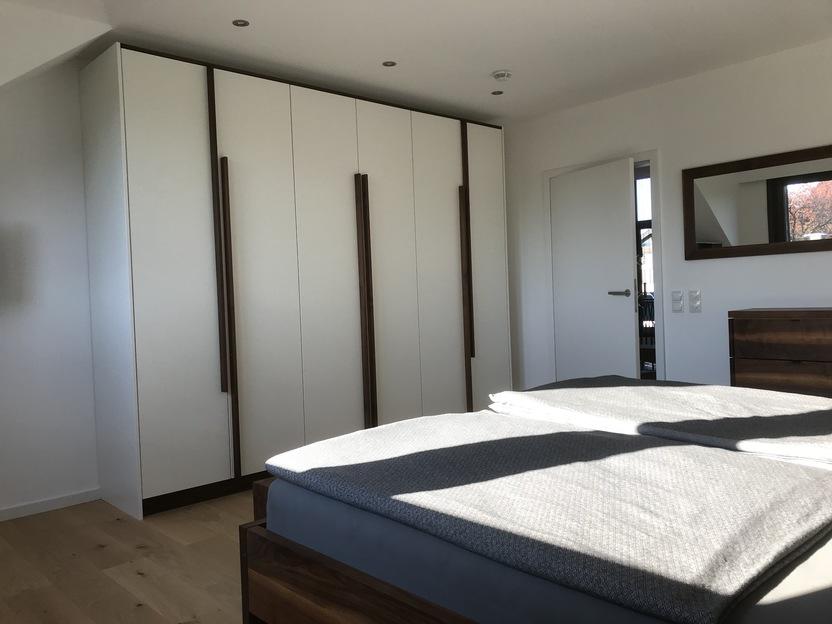 Helles Schlafzimmer mit großem weißen Schrank, Holzdetails und einem Bett mit grauer Bettwäsche.